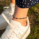 Ръчно изработена гривна за глезен с цветни мъниста, съчетана с бели спортни обувки на зелена трева.