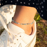 Гривна за глезен с цветни мъниста, съчетана с бяла спортна обувка на фона на тревиста настилка.