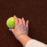 Ръка, държаща тенис топка на червена настилка, с елегантна тенис гривна.