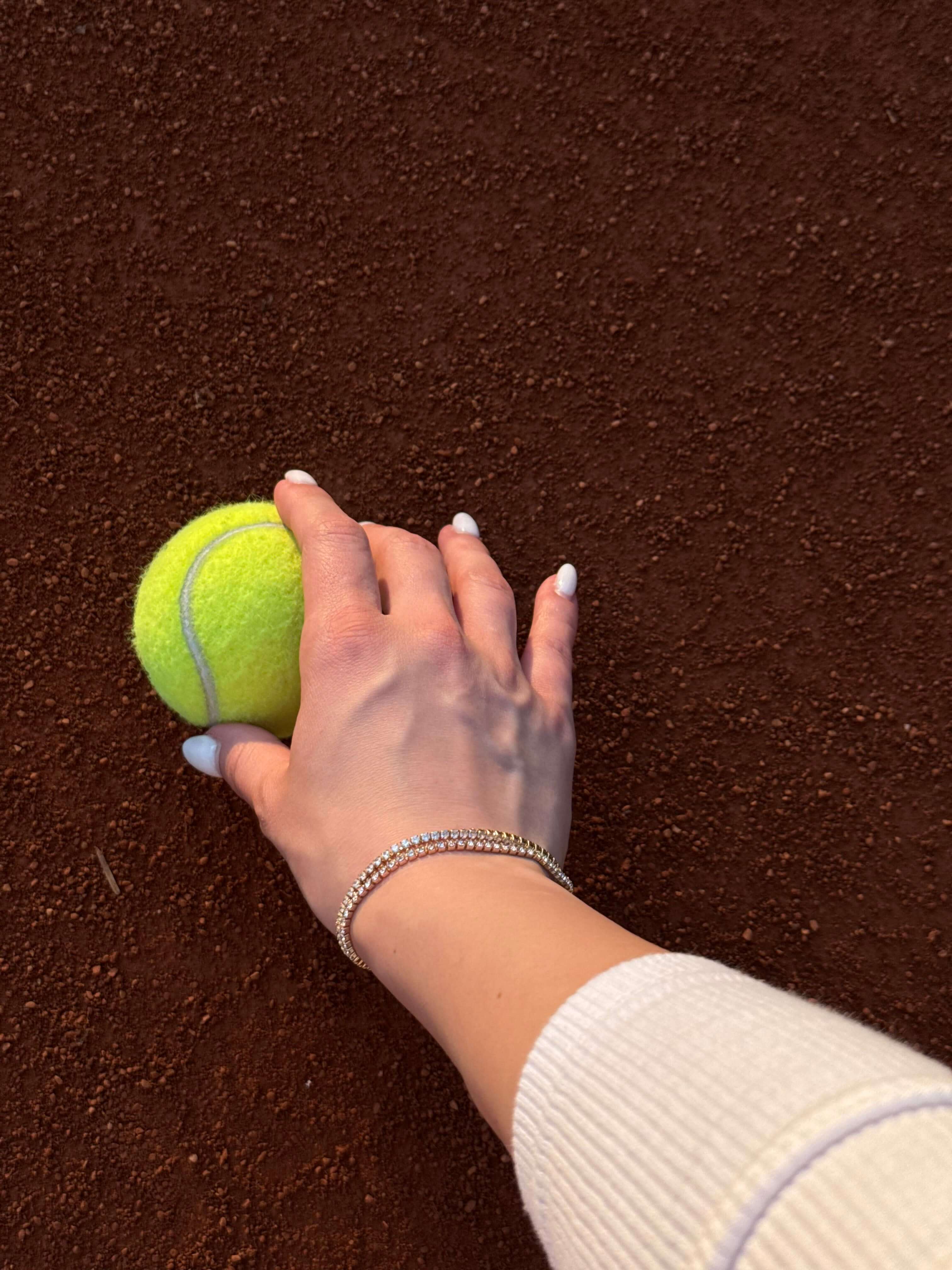 Ръка, държаща тенис топка на червена настилка, с елегантна тенис гривна.