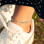 Гривна за глезен с цветни мъниста, съчетана с бяла спортна обувка на фона на тревиста настилка.