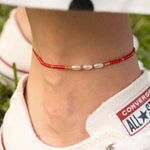 Бохо гривна за глезен от червен конец с перлички, носена с удобни маратонки на зелен фон.