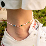 Бохо гривна за глезен с цветни мъниста, носена с летни обувки на тревна повърхност.