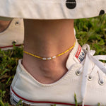 Бохо гривна за глезен с жълти мъниста и перли, носена с бели кеди на зелена трева.