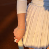 Ръка с тенис ракета, носеща елегантна бяла пола и тънка гривна на тенис корта.