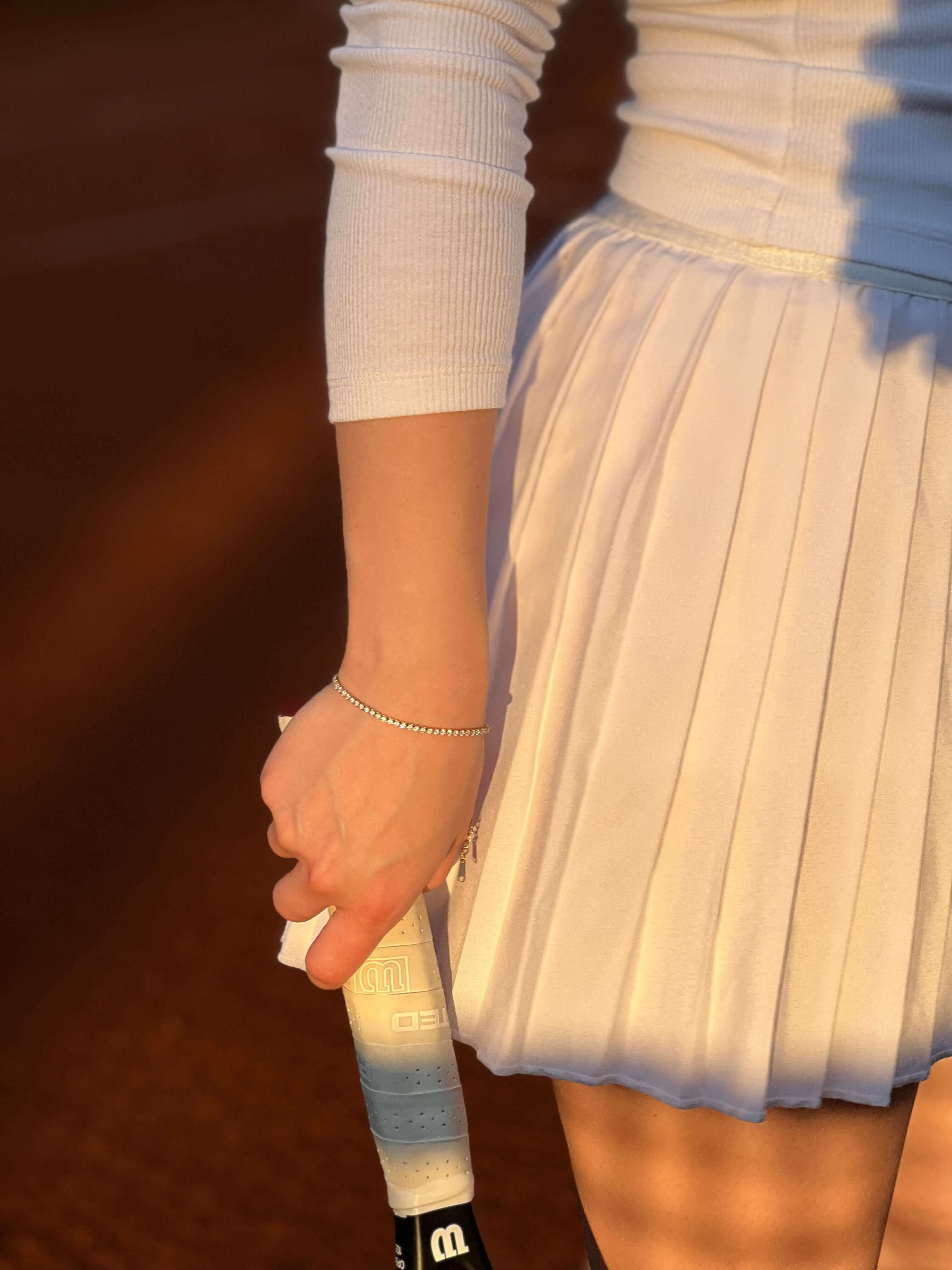 Ръка с тенис ракета, носеща елегантна бяла пола и тънка гривна на тенис корта.