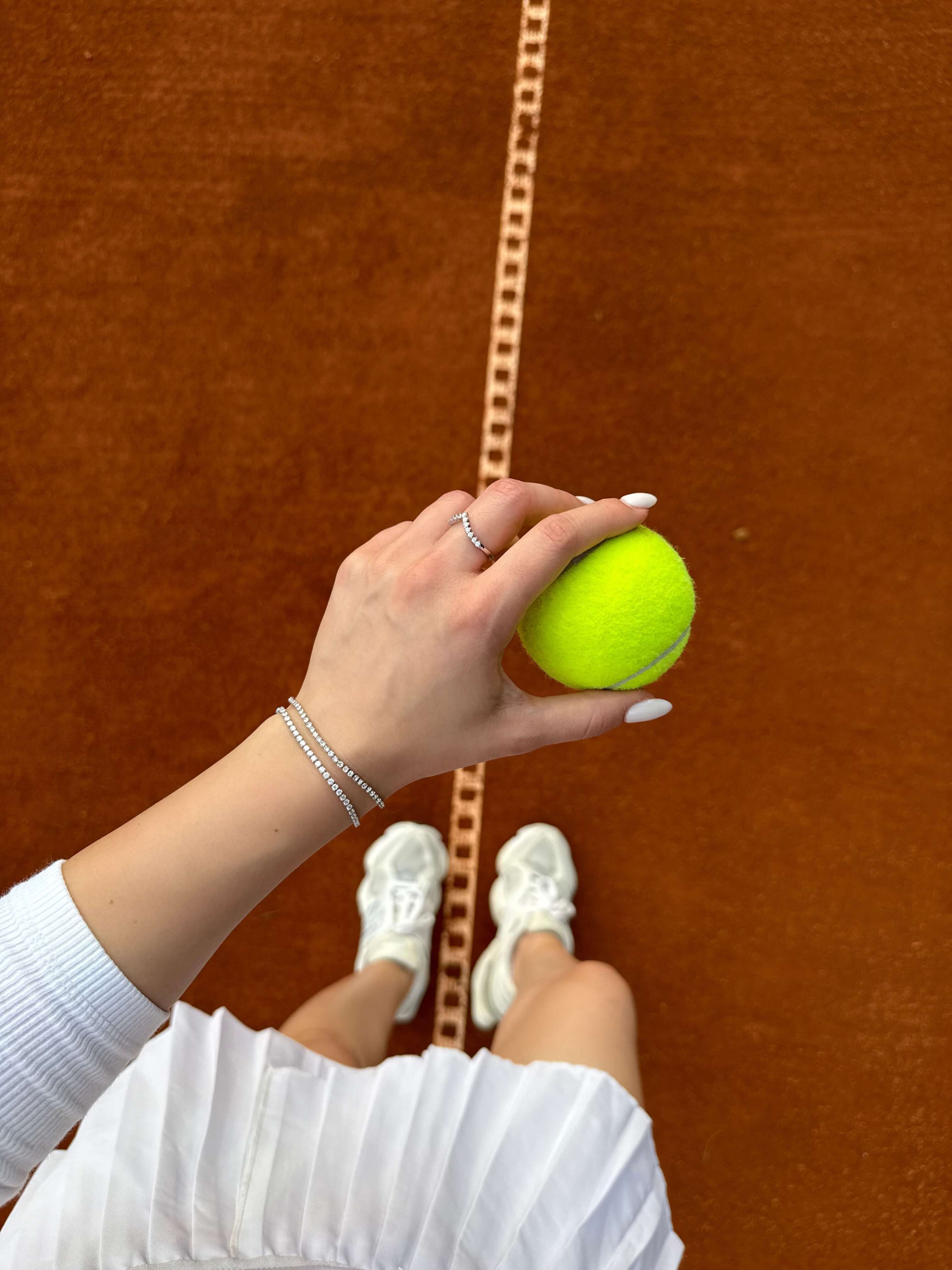 Ръка с тенис топка на тенис корт, носеща стилна гривна на фона на оранжево покритие.