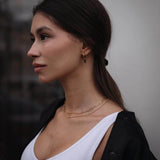 Млада жена с двойно златисто колие и обеци, подчертаваща елегантен стил на фона на неясен градски пейзаж.