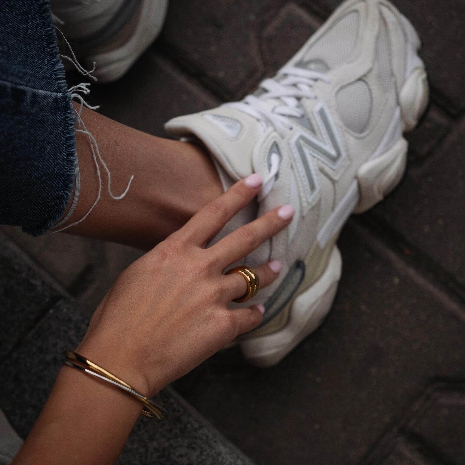 Ръка с златен пръстен и гривна, поставена до бяла маратонка New Balance на улицата.