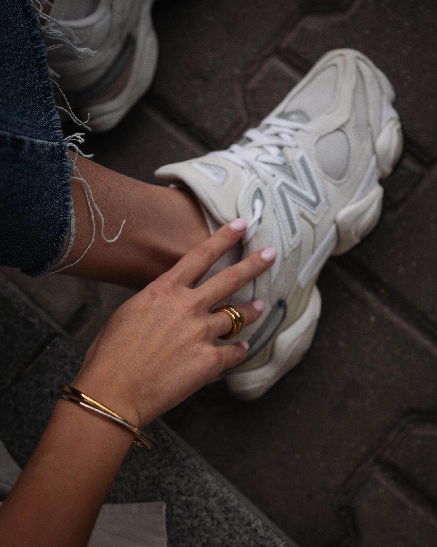 Ръка с златен пръстен и гривна, поставена до бяла маратонка New Balance на улицата.