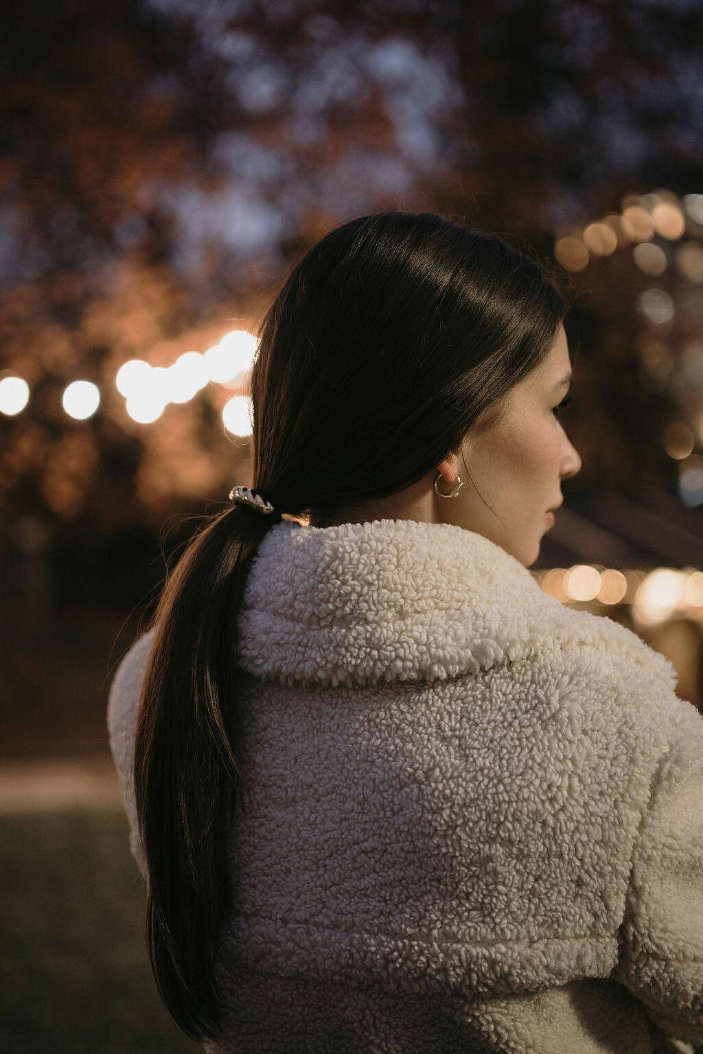 Жена с дълга коса, вързана в опашка, облечена в уютно палто на фона на вечерни светлини.