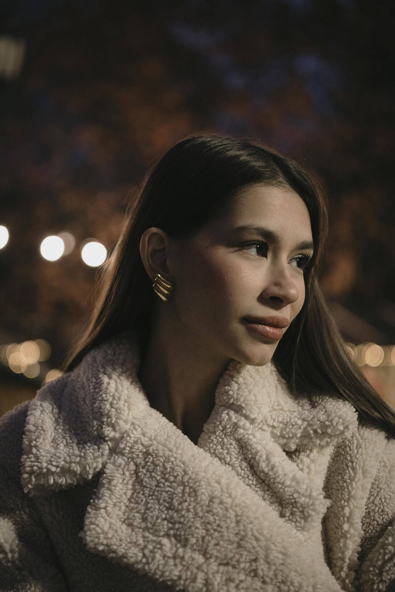 Млада жена с обеци "Криле" с 18К златно покритие, облечена в пухкаво палто, на фона на вечерна светлина.