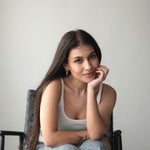 Young woman sitting in a chair with long hair, wearing a casual outfit, smiling thoughtfully at the camera.