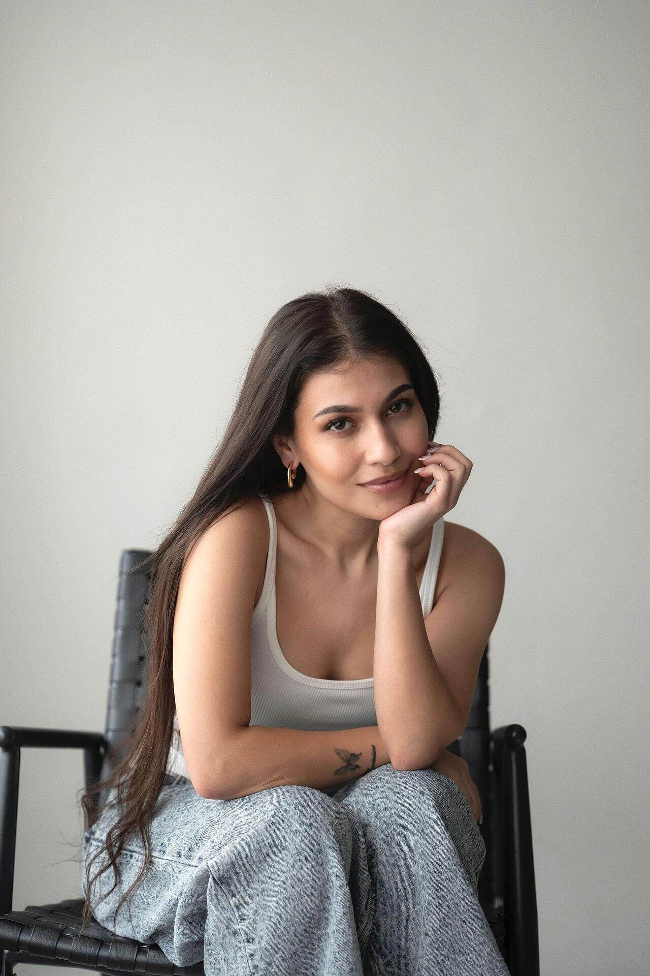 Young woman sitting in a chair with long hair, wearing a casual outfit, smiling thoughtfully at the camera.