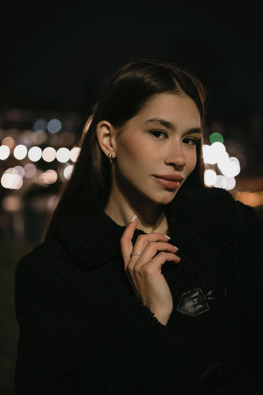 Модерна жена с обеци V-Shape, облечена в черно палто на фона на нощен град с размазани светлини.