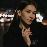 Модерна жена с обеци V-Shape, облечена в черно палто на фона на нощен град с размазани светлини.