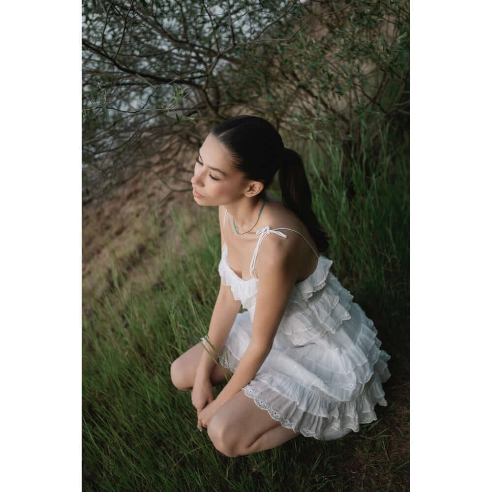 Млада жена с дълга коса седнала в зелена трева, носеща бяла рокля с ръкави, в естествена среда.