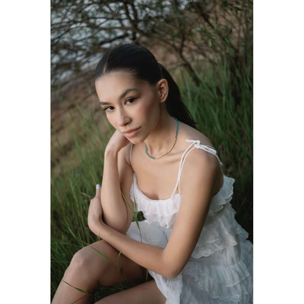 Модел носи колие Wave Син Тюркоаз с 18К позлата на фона на природа, облечен в бяла рокля.