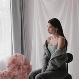 Млада жена в сива жилетка с огледален вид, седнала пред розово сърце и завеси.