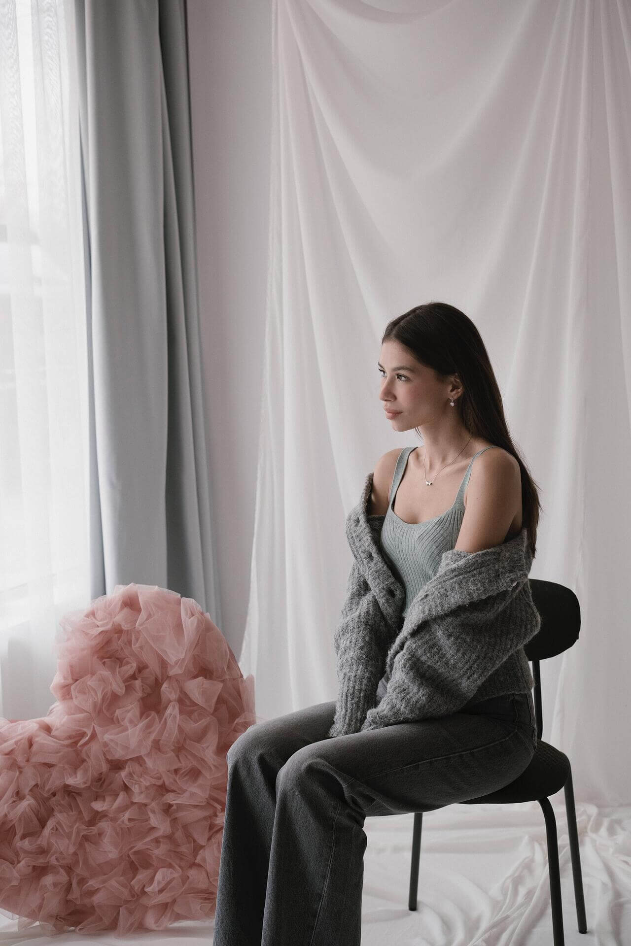 Млада жена в сива жилетка с огледален вид, седнала пред розово сърце и завеси.