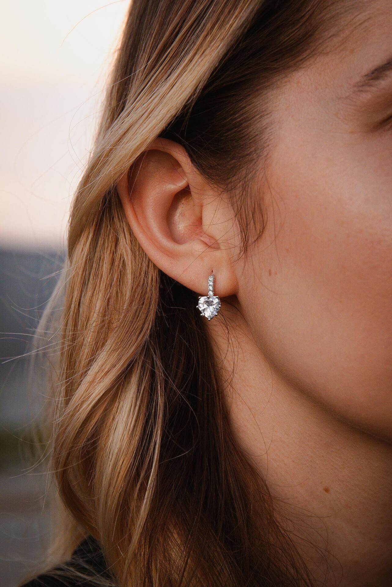 Елегантни сребърни обеци с форма на сърце, носени от жена с дълга коса на фон на залез.