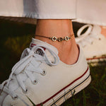 Гривна за глезен от медицинска стомана в златист цвят, носена с бели маратонки на фон от зелена трева.