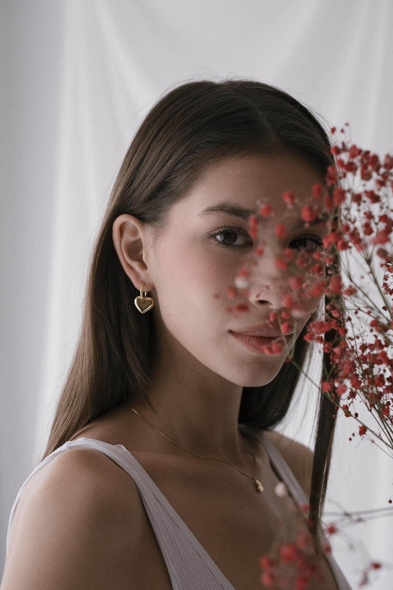 Жена с обеци сърце от 18K златно покритие, зад която се виждат червени цветя.