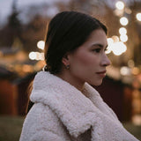 Млада жена в бяла палто с елегантни обеци, на фона на вечерни светлини.