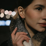 Млада жена с златисти обеци с многонюансни кристали, облечена в тъмен шал, на фона на размазани светлини.