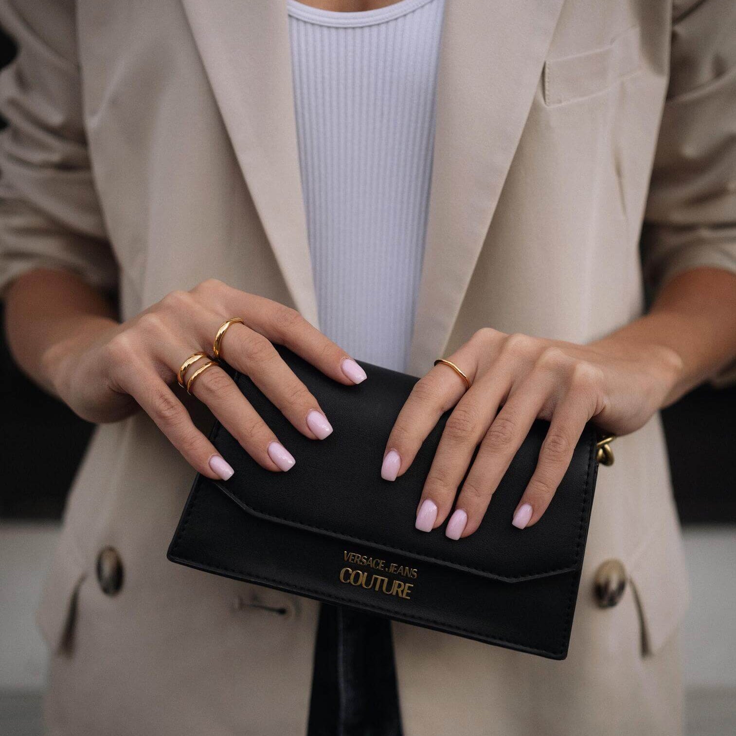 Жена с изискано облекло държи черен портфейл, носейки златисти пръстени и нежно маникюр.